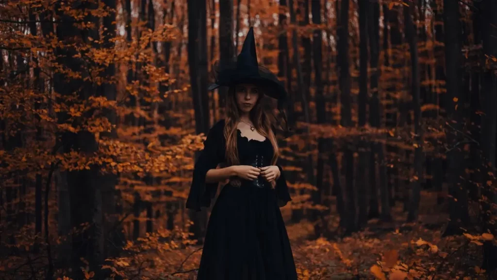 Photo of a woman dressed as a witch in a dense fall forest. She poses in the center of the image. Themes of 2024 Player's Handbook.